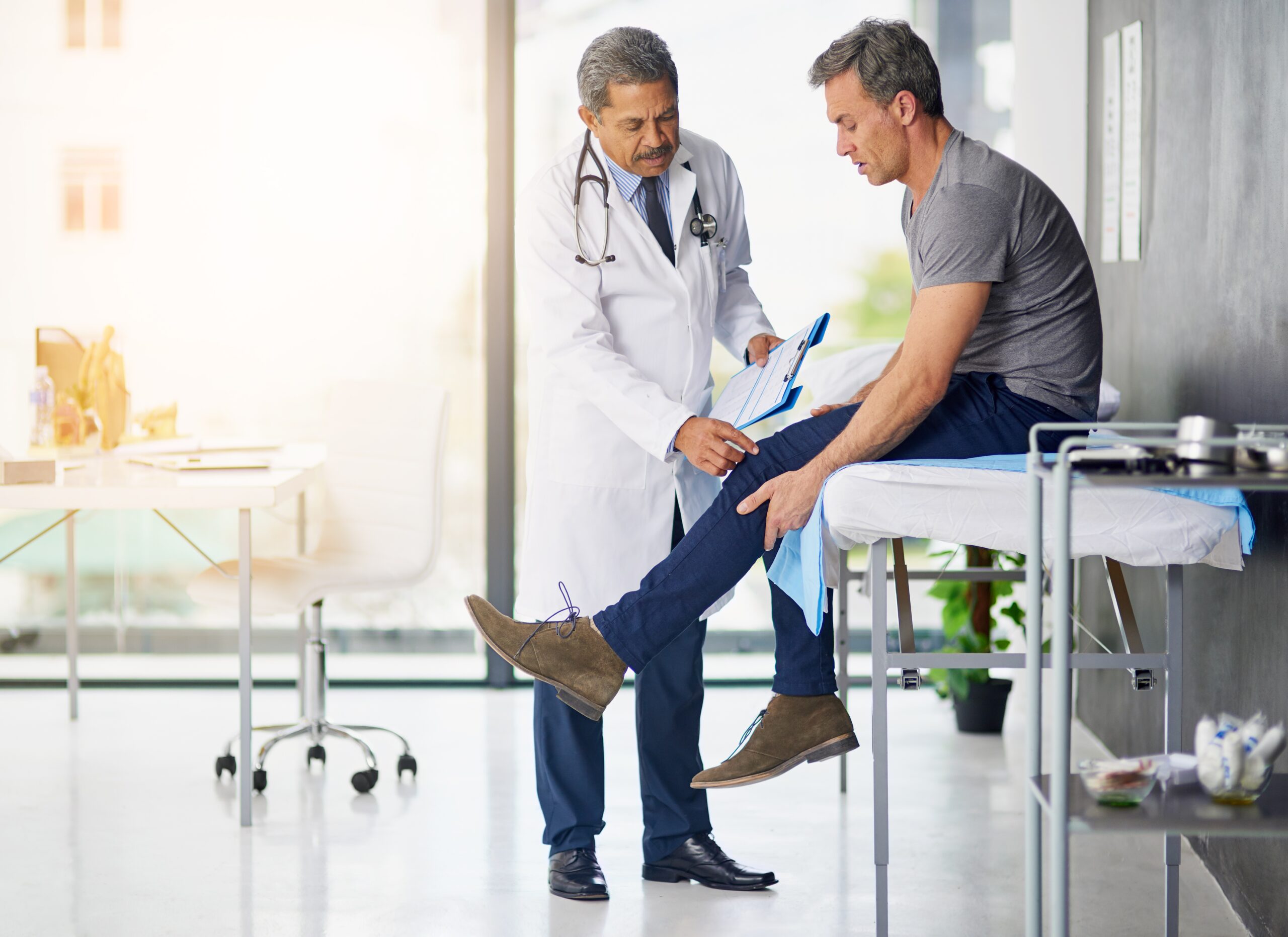 A doctor in a white coat examines a male patient’s leg as he sits on a medical table in a modern, well-lit clinic - Alan Dayan, MD, PC, FAAOS A doctor in a white coat examines a male patient’s leg as he sits on a medical table in a modern, well-lit clinic - Alan Dayan, MD, PC, FAAOS